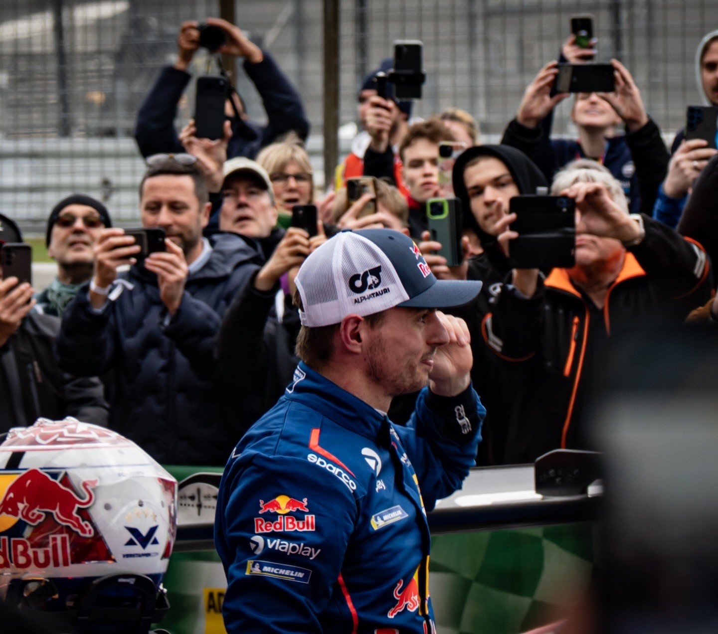 Rennfahrer im Red-Bull-Rennanzug bei einer Motorsport-Veranstaltung, umringt von Fans mit Smartphones - Fotografie von Nik Pottbecker